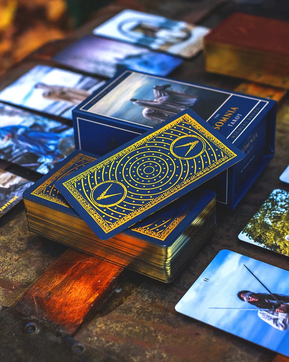 Nicolas Bruno's Somnia Tarot card back resting on its angled box on a rustic wooden table, surrounding cards softly blurred