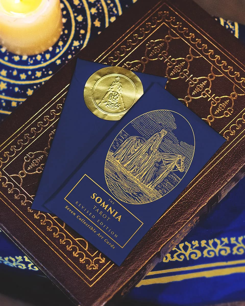 Somnia Tarot art card envelope with a gold wax seal and engraved rider motif, resting on a gilt-tooled leather book by candlelight