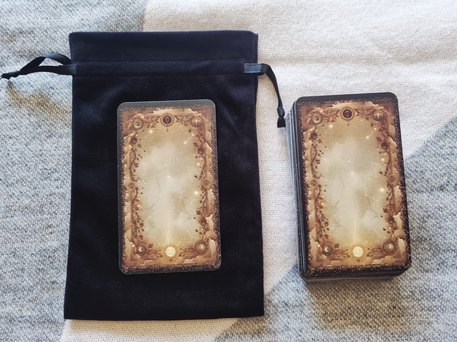 Overhead view of a black velvet drawstring bag and stacked tarot deck with gilded card backs, on a woven cloth