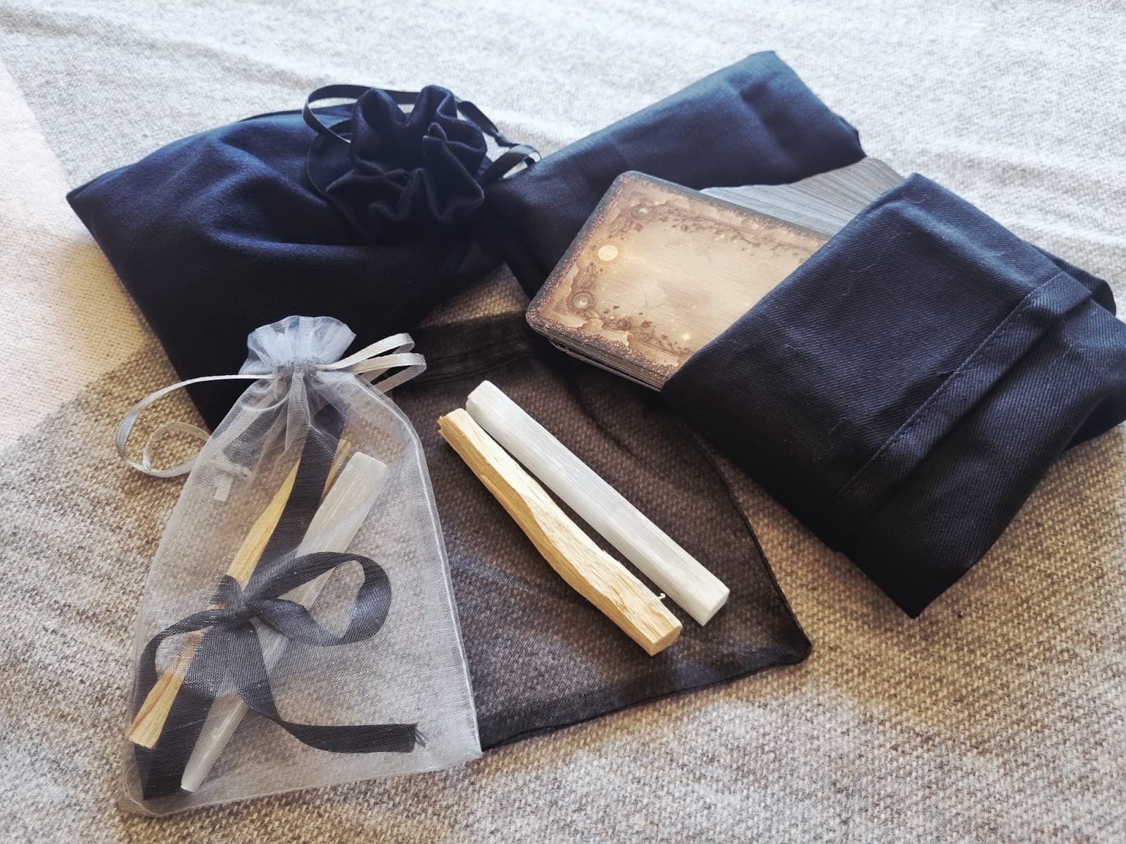 Black velvet tarot bag, folded cloth and card deck pictured alongside a palo santo bundle and organza gift bag on a beige throw